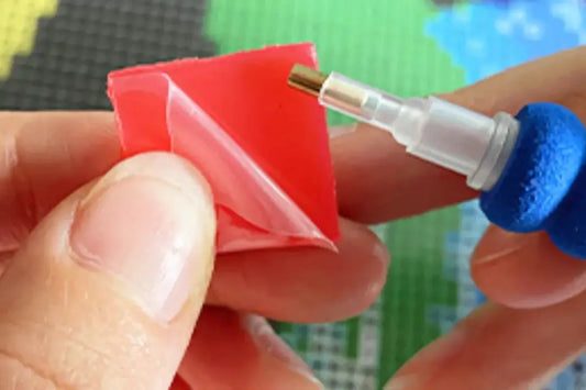 Close-up van een hand die roze wax afpelt voor een diamond painting pen, met een kleurrijke diamond painting ondergrond op de achtergrond.