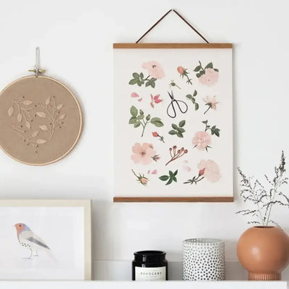 Moderne interieurdecoratie met een houten posterhanger die een kunstafdruk met bloemenmotieven presenteert, naast een borduurhoepel en decoratieve items op een witte muur.