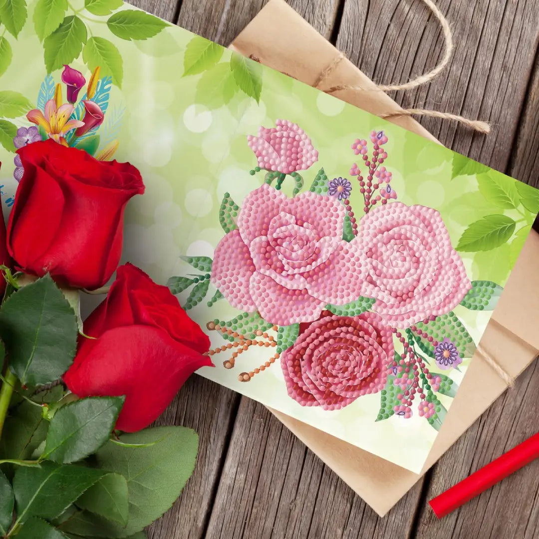 Diamond painting wenskaart met roze rozen en groene bladeren op een houten tafel.