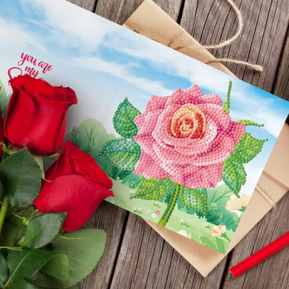 Diamond painting wenskaart met een grote roze roos en rode rozen op een houten tafel.