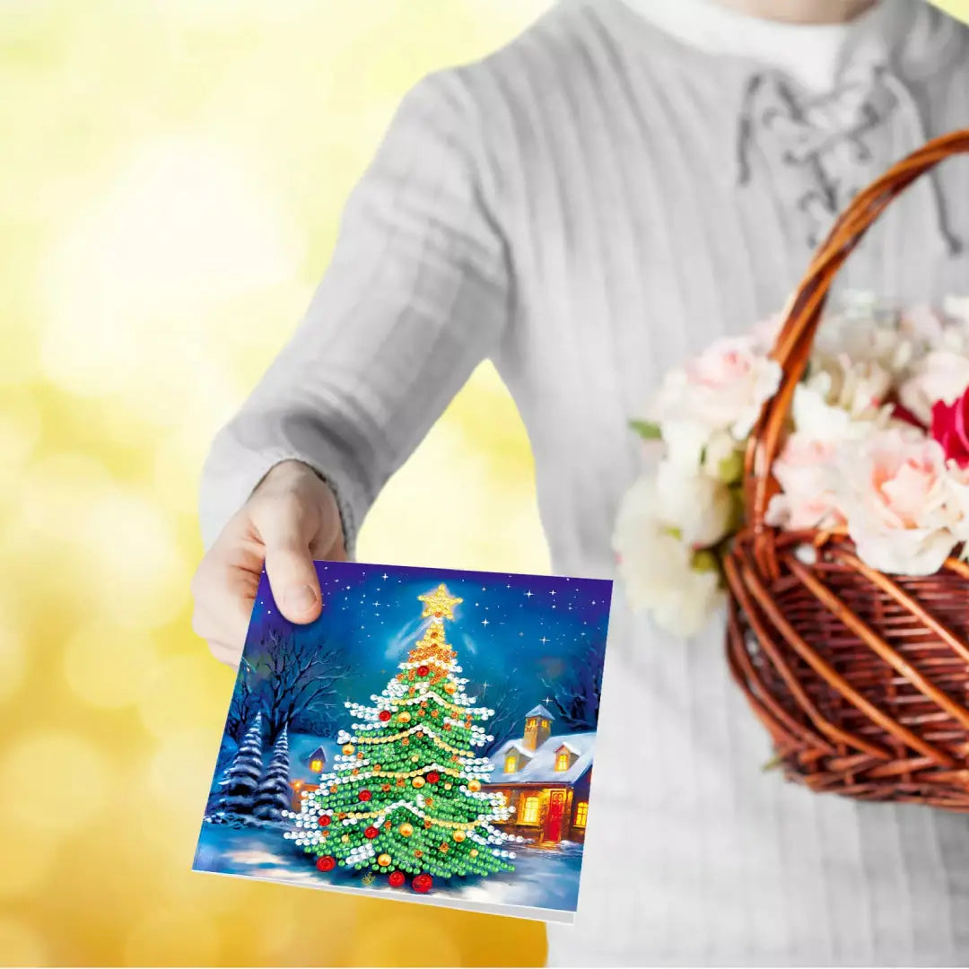 Vrouw houdt een diamond painting kerstkaart met een feestelijk verlichte kerstboom in een sneeuwlandschap vast