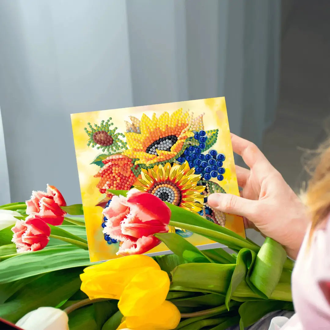 Vrouwelijke handen plaatsen een diamond painting kaart met zonnebloemen en druiven tegen een achtergrond van tulpen, een mix van kunst en natuur.