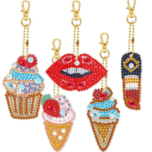 Collectie van diamond painting sleutelhangers in de vorm van cupcakes, ijsjes, en rode lippen, rijk gedecoreerd met kleurrijke steentjes.