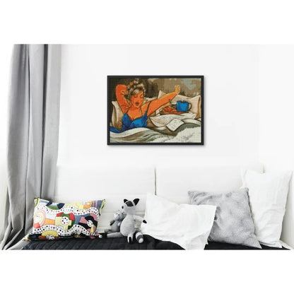 Ingelijst diamond painting van een vrouw in bed met een boek en een kopje koffie, opgehangen boven een wit bed met kleurrijke kussens.