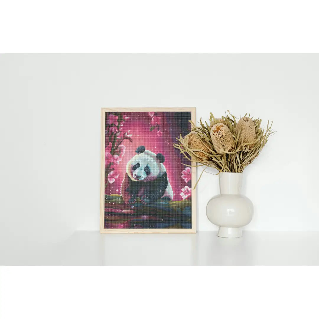 Diamond painting van een panda met een roze achtergrond en bloemen, geplaatst naast een witte vaas met droogbloemen.